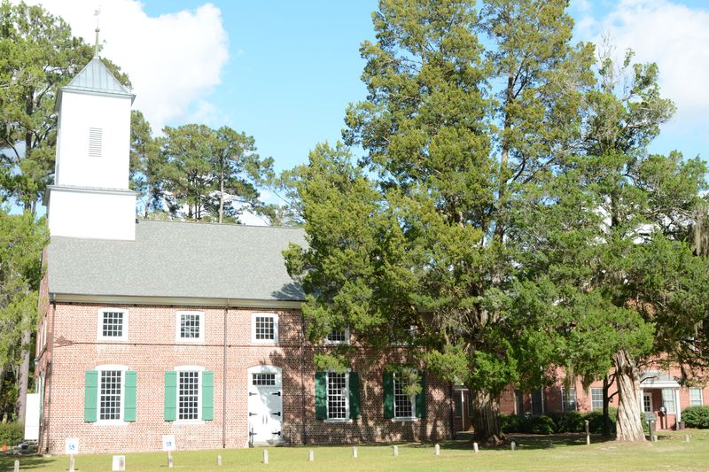 Long side of the brick church with green shutters, the white steeple at left and a large tree at right