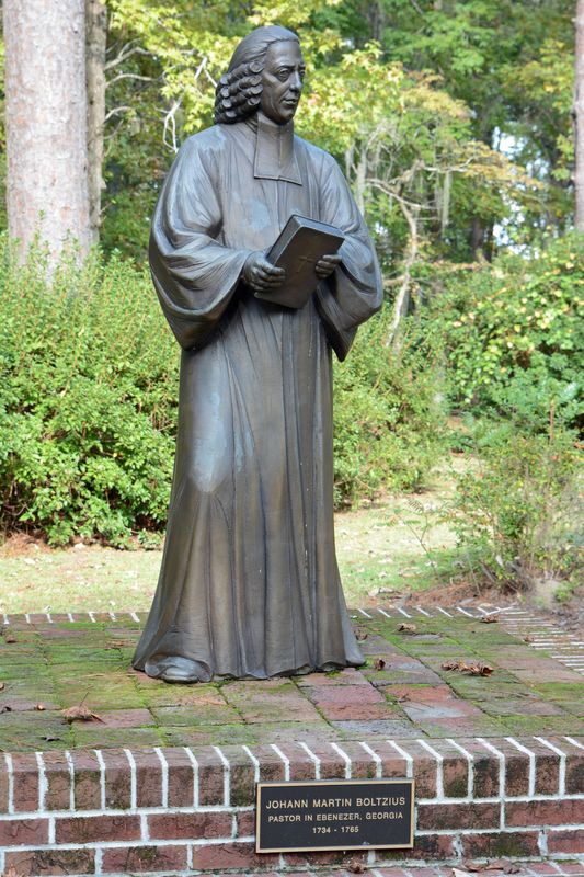 Bronze statue of Pastor Boltzius holding a book, on a brick base among trees